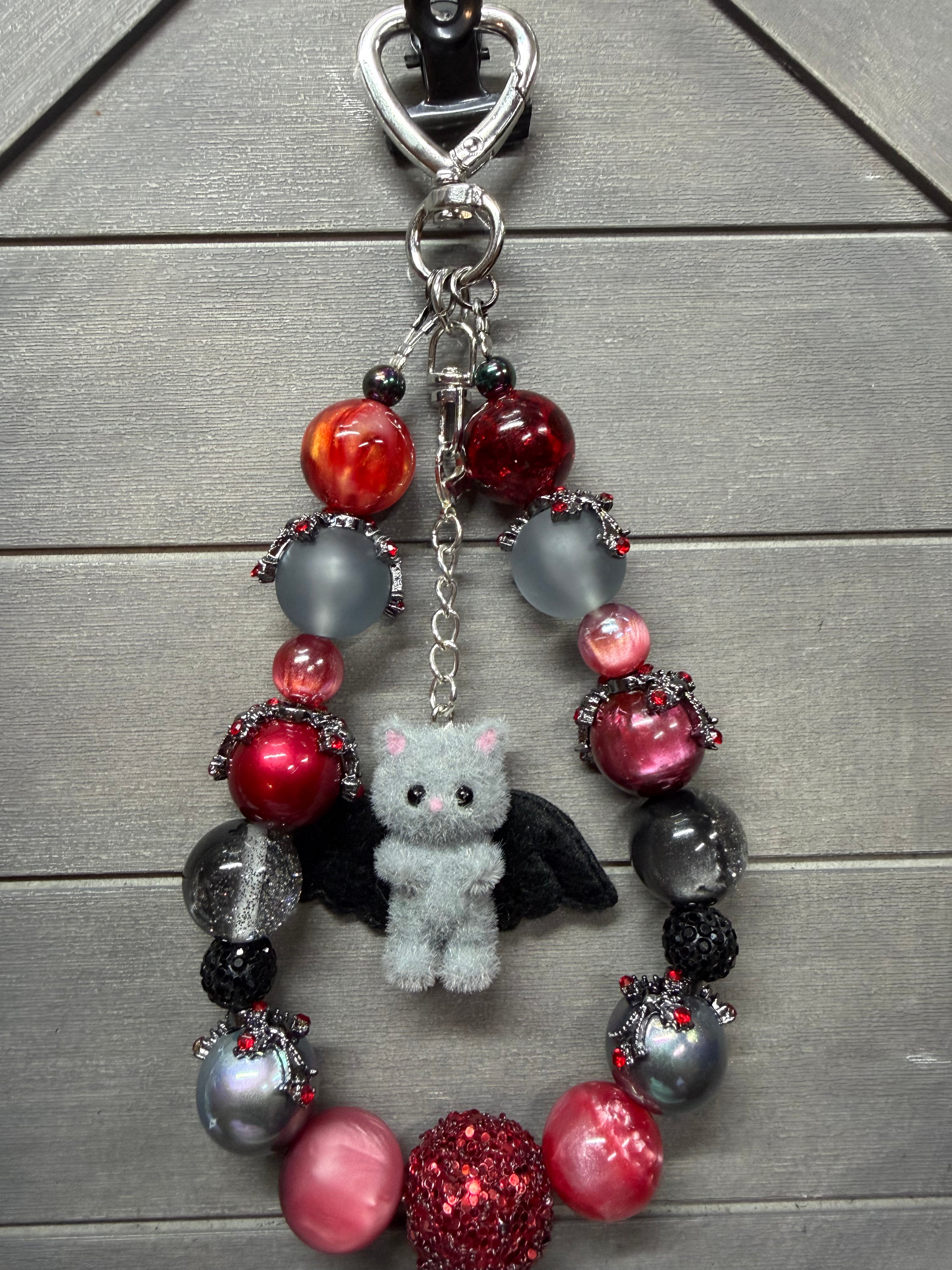 A beaded charm featuring red, pink, black, and gray beads, a small gray plush bunny with black wings, and a silver heart-shaped clasp, displayed on a gray wood plank background.