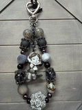 A decorative bag charm with a silver clasp, featuring matte black beads, faceted glass beads, a small anime-style character charm, and a silver rose bead on a gray wood background.