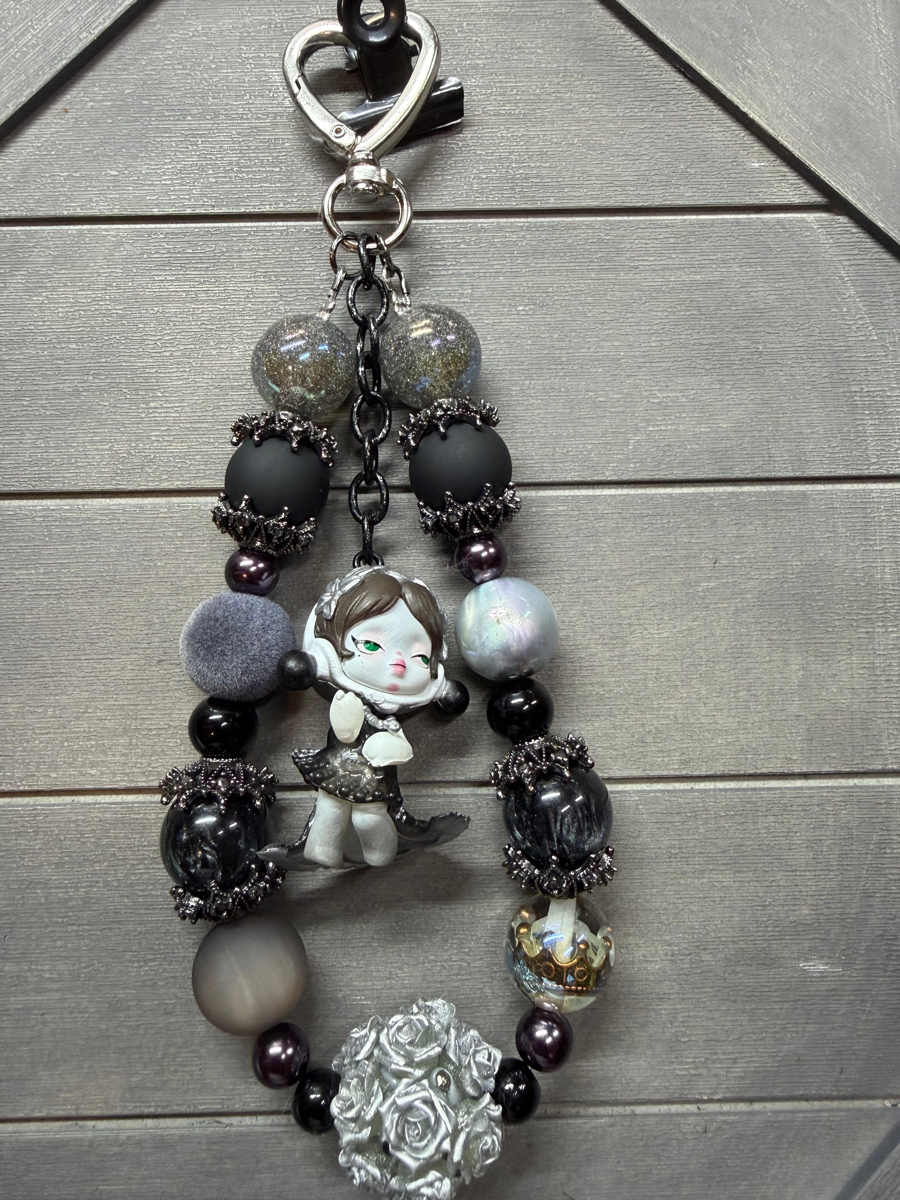 A decorative bag charm with a silver clasp, featuring matte black beads, faceted glass beads, a small anime-style character charm, and a silver rose bead on a gray wood background.