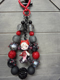 A gothic-themed beaded charm featuring a small anime doll charm with red hair, surrounded by matte black, metallic silver, and red rhinestone beads, displayed on a gray wood background.