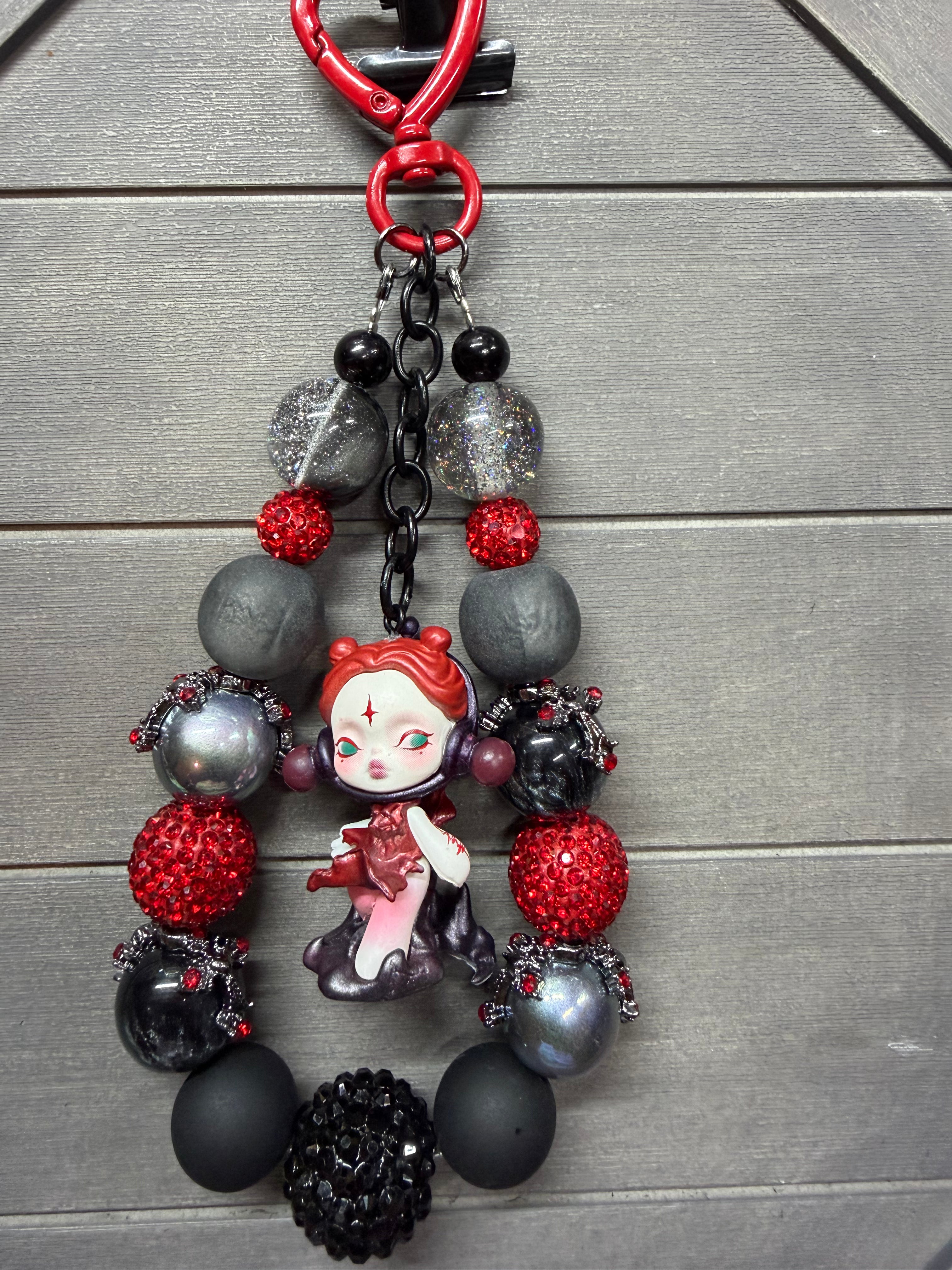 A gothic-themed beaded charm featuring a small anime doll charm with red hair, surrounded by matte black, metallic silver, and red rhinestone beads, displayed on a gray wood background.