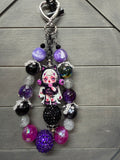 A gothic-themed charm featuring a small anime doll charm with pink eyes and a white dress, surrounded by purple, black, and silver beads, on a gray wood background.