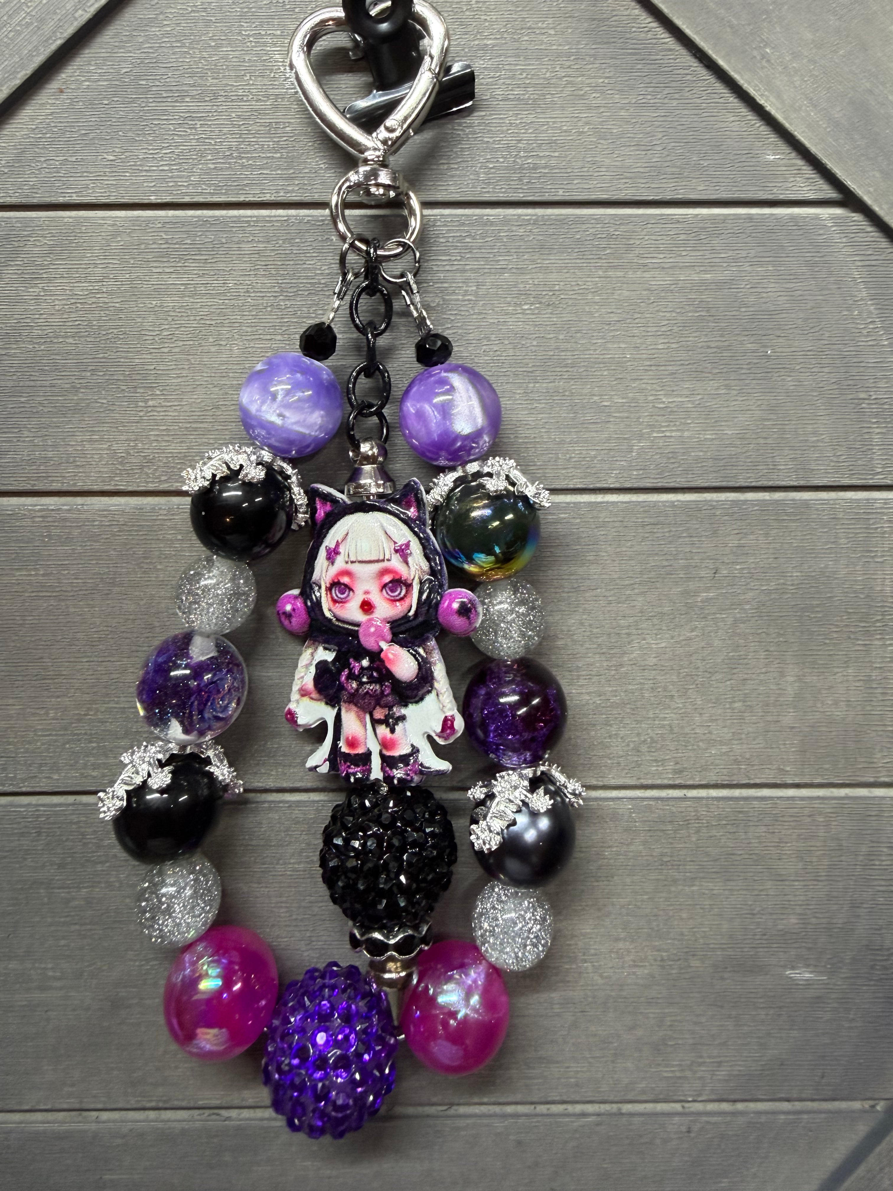 A gothic-themed charm featuring a small anime doll charm with pink eyes and a white dress, surrounded by purple, black, and silver beads, on a gray wood background.
