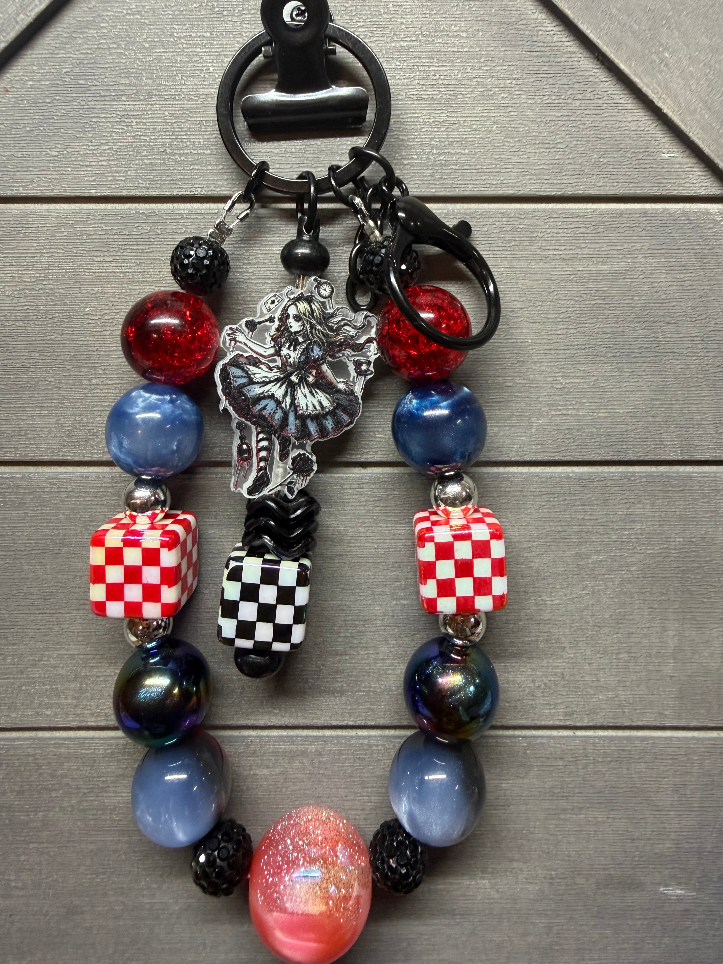 A beaded charm with a black metal split ring and clasp, featuring red and blue glass beads, checkered cube beads, a silver key charm, and a black-and-white checkerboard tassel, displayed on a gray wood background.