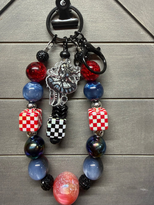 A beaded charm with a black metal split ring and clasp, featuring red and blue glass beads, checkered cube beads, a silver key charm, and a black-and-white checkerboard tassel, displayed on a gray wood background.