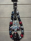 A gothic-themed beaded keychain featuring a central Alice in Wonderland illustration charm, black beads, and red and blue rhinestone accents, shown on a gray wood plank background.