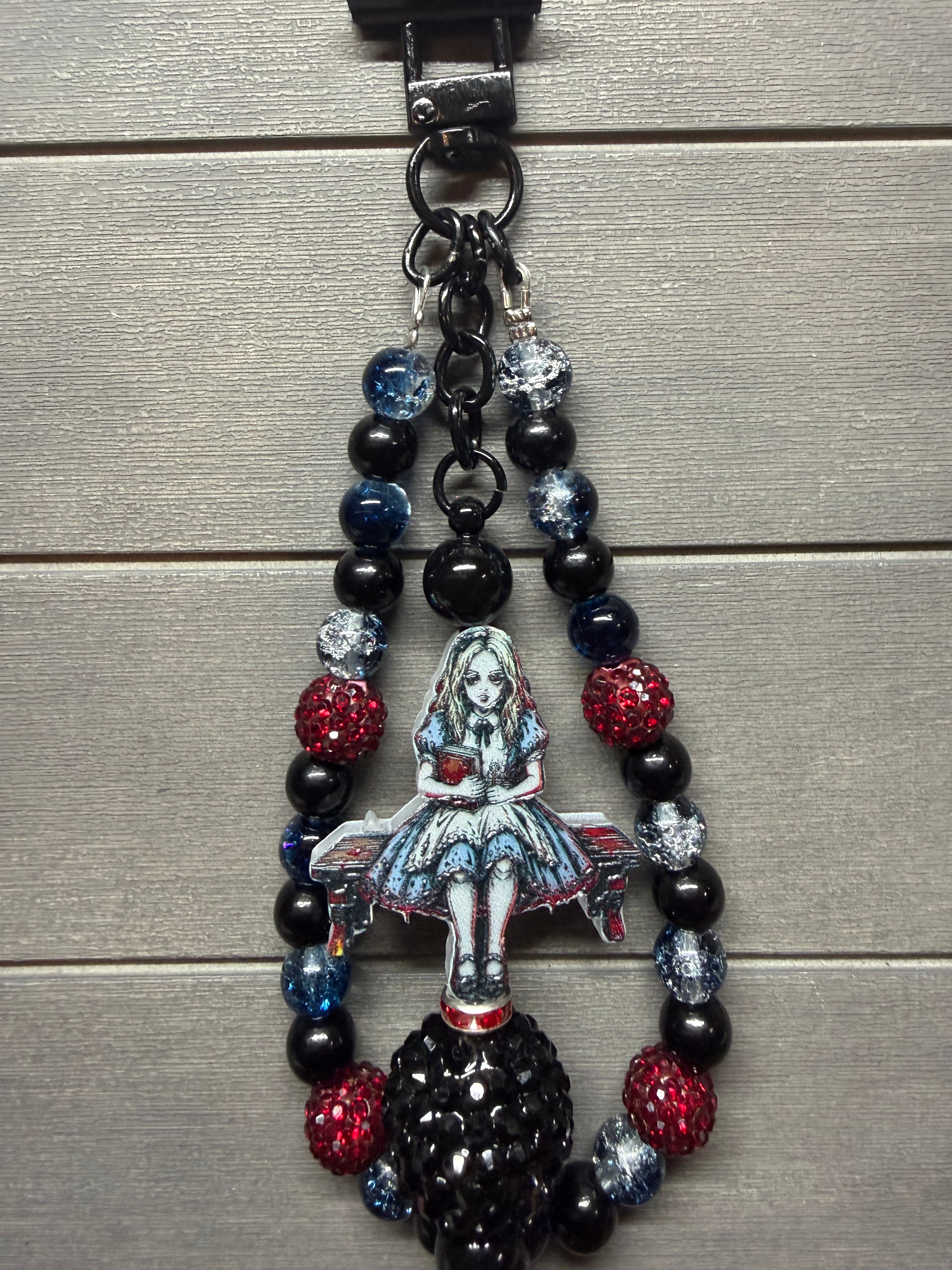 A gothic-themed beaded keychain featuring a central Alice in Wonderland illustration charm, black beads, and red and blue rhinestone accents, shown on a gray wood plank background.