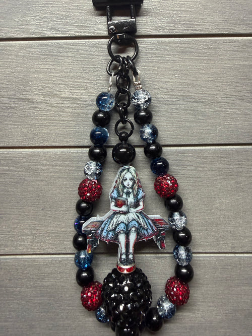 A gothic-themed beaded keychain featuring a central Alice in Wonderland illustration charm, black beads, and red and blue rhinestone accents, shown on a gray wood plank background.