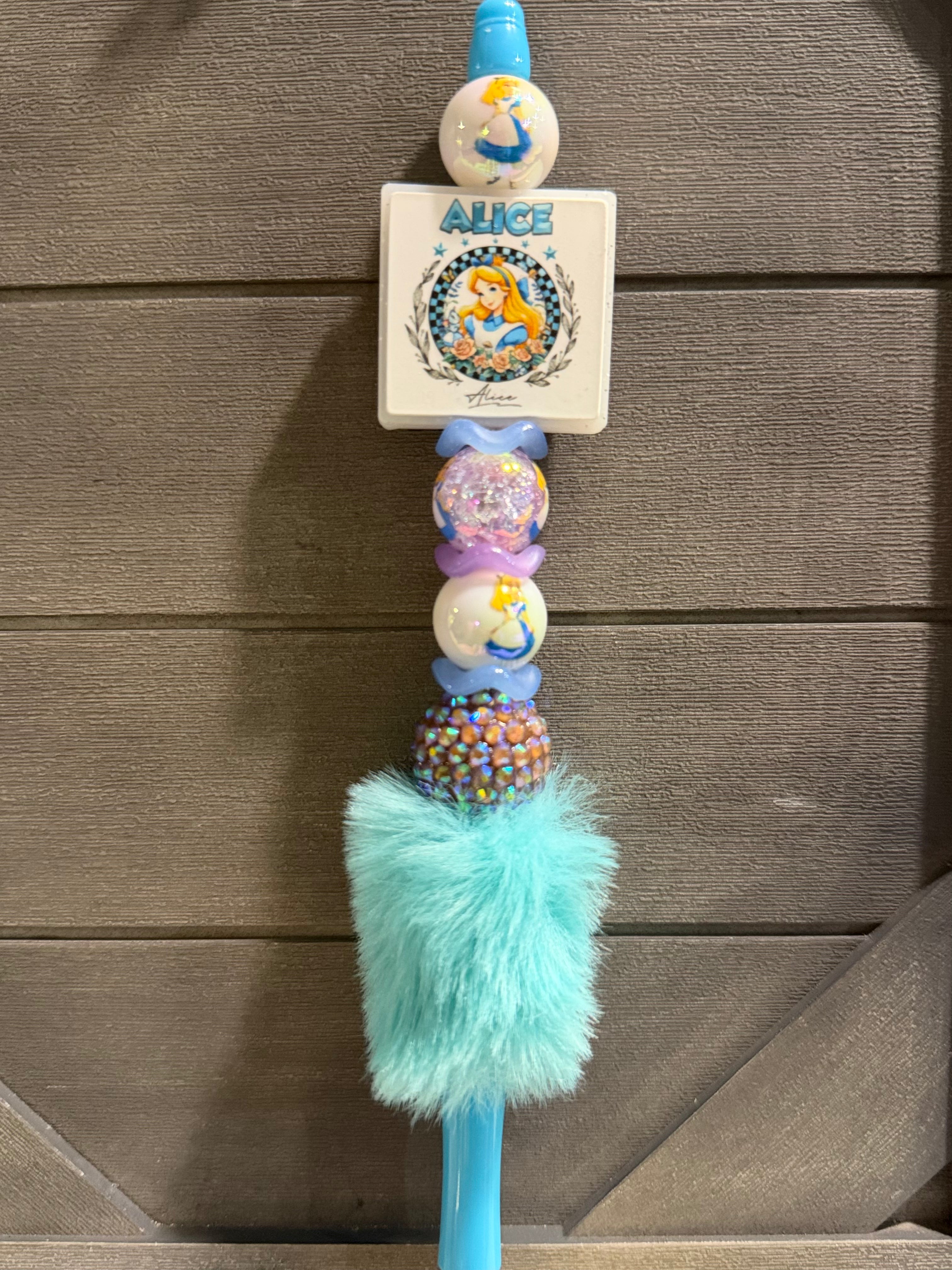 A personalized beaded wand or keychain featuring a white square card with 'ALICE' and a cartoon illustration, decorated with blue, purple, and multicolor beads, a fluffy mint green pom-pom, and a blue plastic end, shown against a brown wooden background.
