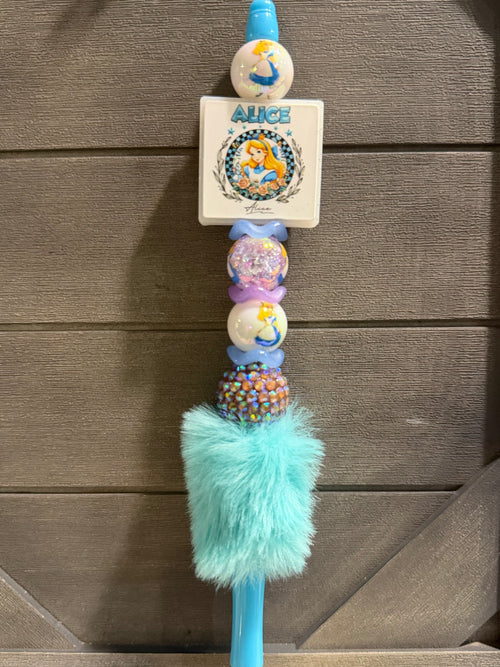 A personalized beaded wand or keychain featuring a white square card with 'ALICE' and a cartoon illustration, decorated with blue, purple, and multicolor beads, a fluffy mint green pom-pom, and a blue plastic end, shown against a brown wooden background.