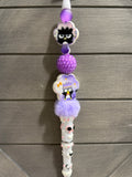 A white pen with purple and lavender charms, including cartoon penguin figures, a textured purple ball, and a fluffy purple pom-pom, shown against a gray wood plank background.