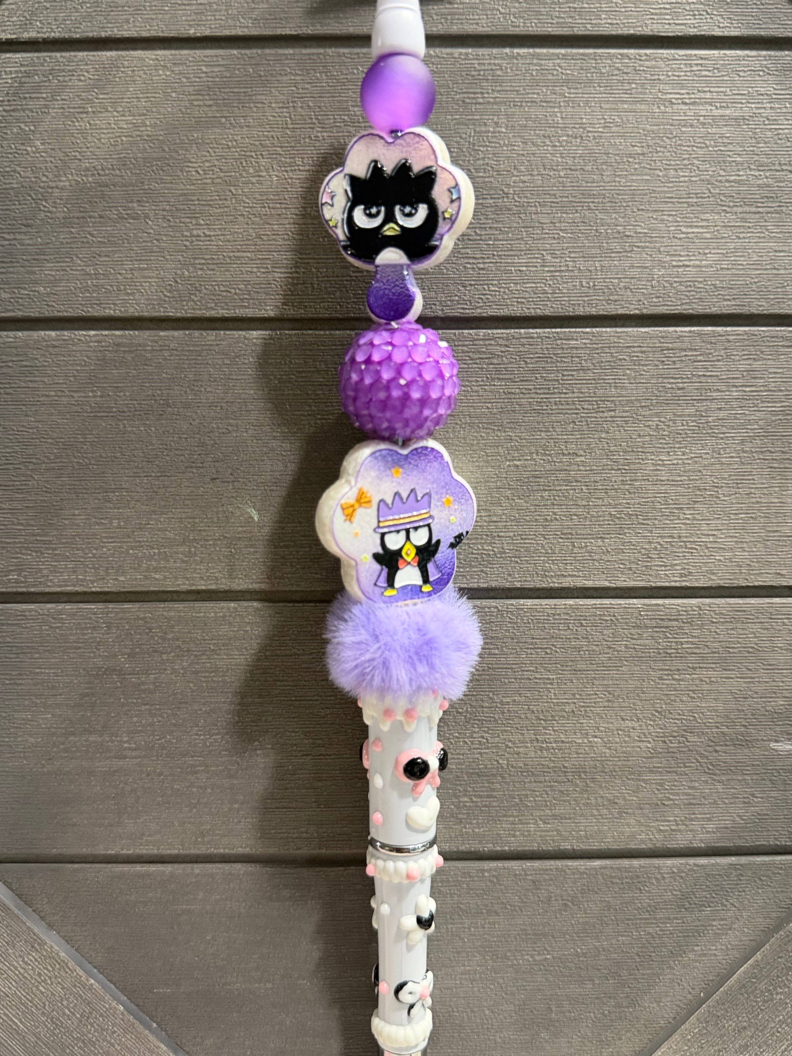 A white pen with purple and lavender charms, including cartoon penguin figures, a textured purple ball, and a fluffy purple pom-pom, shown against a gray wood plank background.