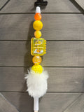 A white plastic pen, decorated with orange and yellow beads, lazy egg, and a white faux fur pom-pom, shown against a gray wood-paneled background.