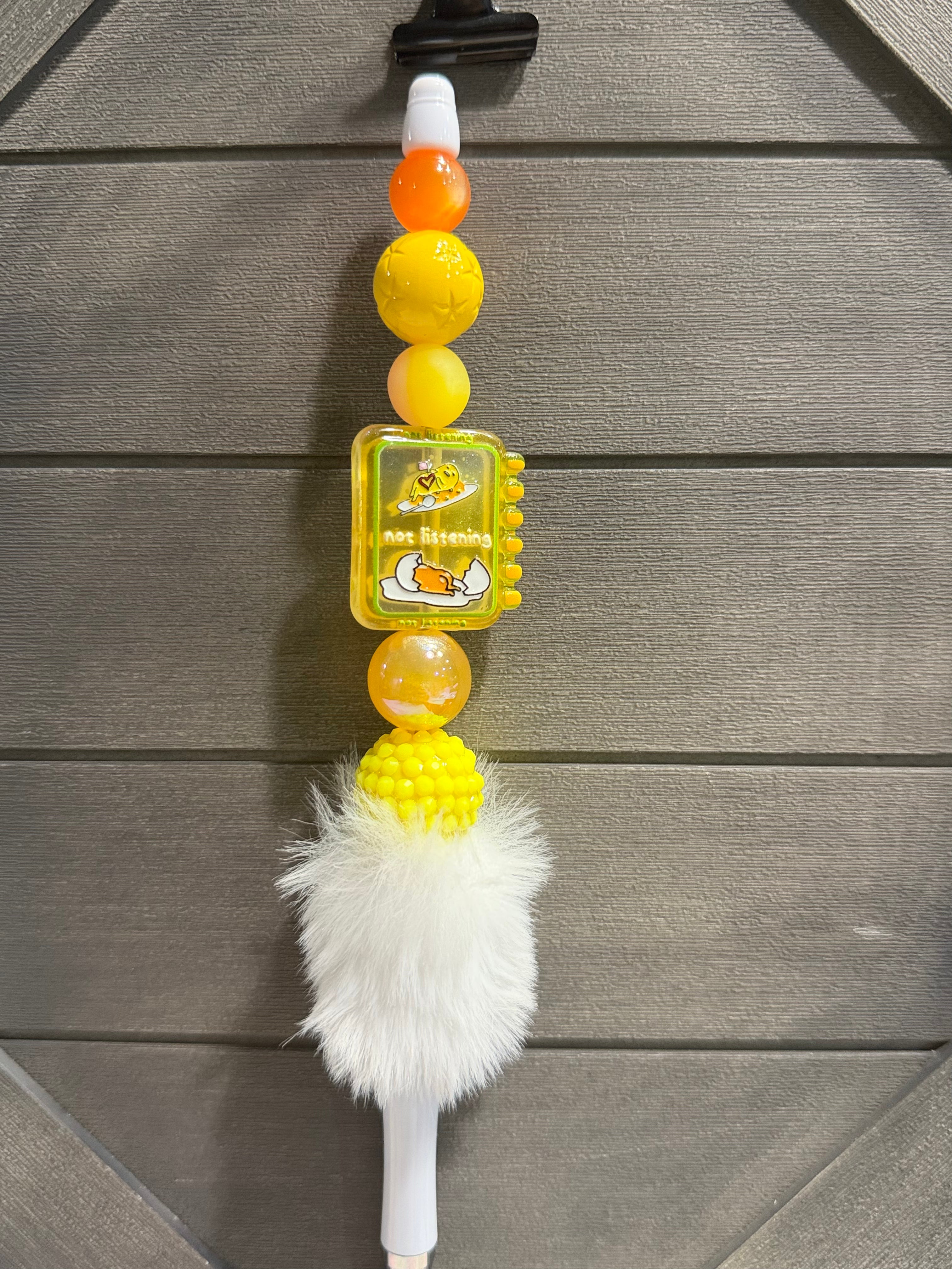 A white plastic pen, decorated with orange and yellow beads, lazy egg, and a white faux fur pom-pom, shown against a gray wood-paneled background.