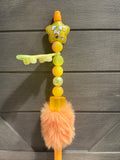 A colorful beaded pen with a yellow cartoon character charm, translucent yellow beads, a fluffy peach pom-pom, and a small key-shaped accent, shown against a dark gray wood plank background.