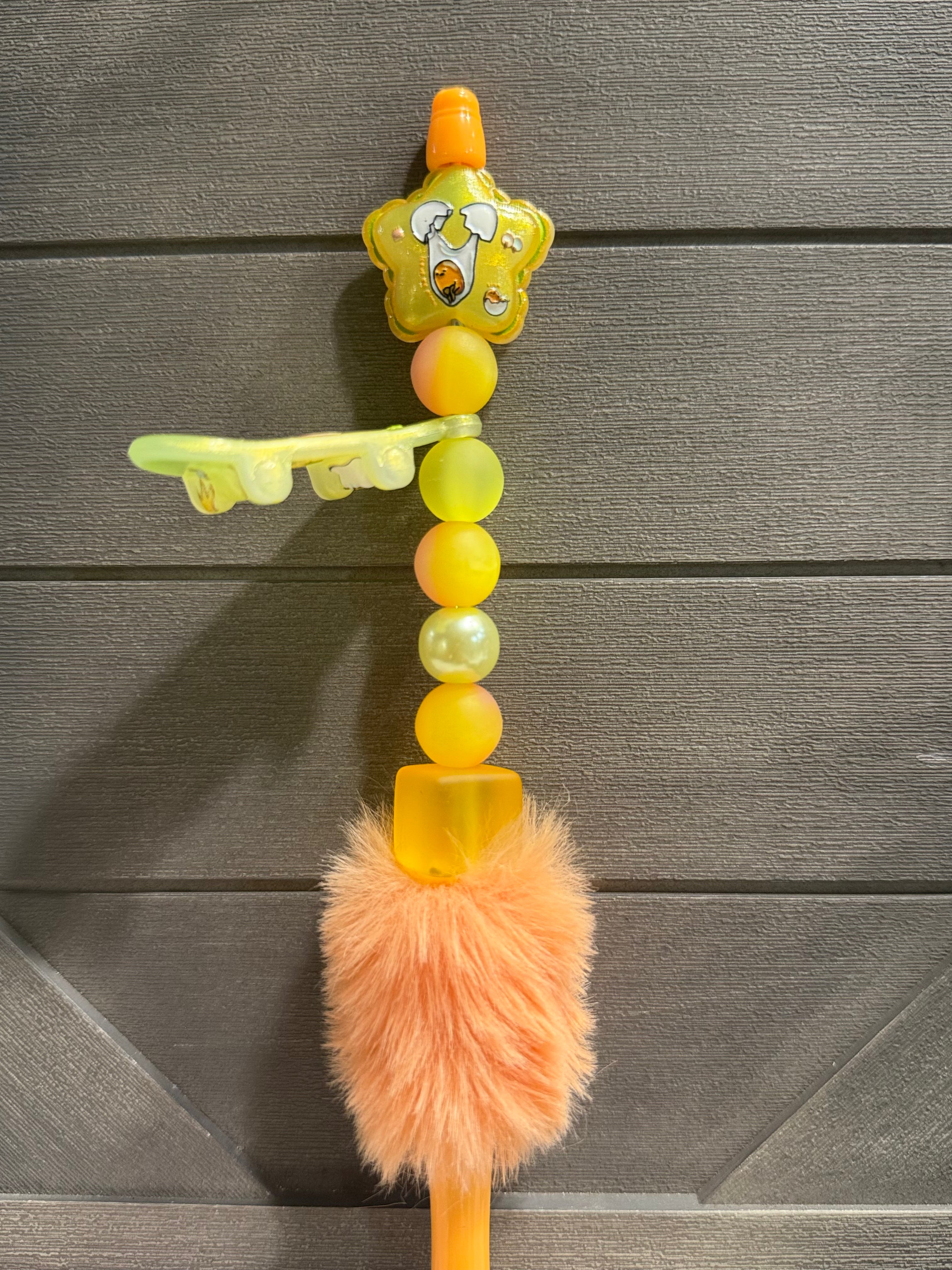 A colorful beaded pen with a yellow cartoon character charm, translucent yellow beads, a fluffy peach pom-pom, and a small key-shaped accent, shown against a dark gray wood plank background.