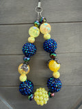 A beaded wristlet with a silver lobster clasp, featuring large iridescent yellow beads, deep blue rhinestone-covered pom-poms, and smaller iridescent beads, displayed on a dark gray wood background.