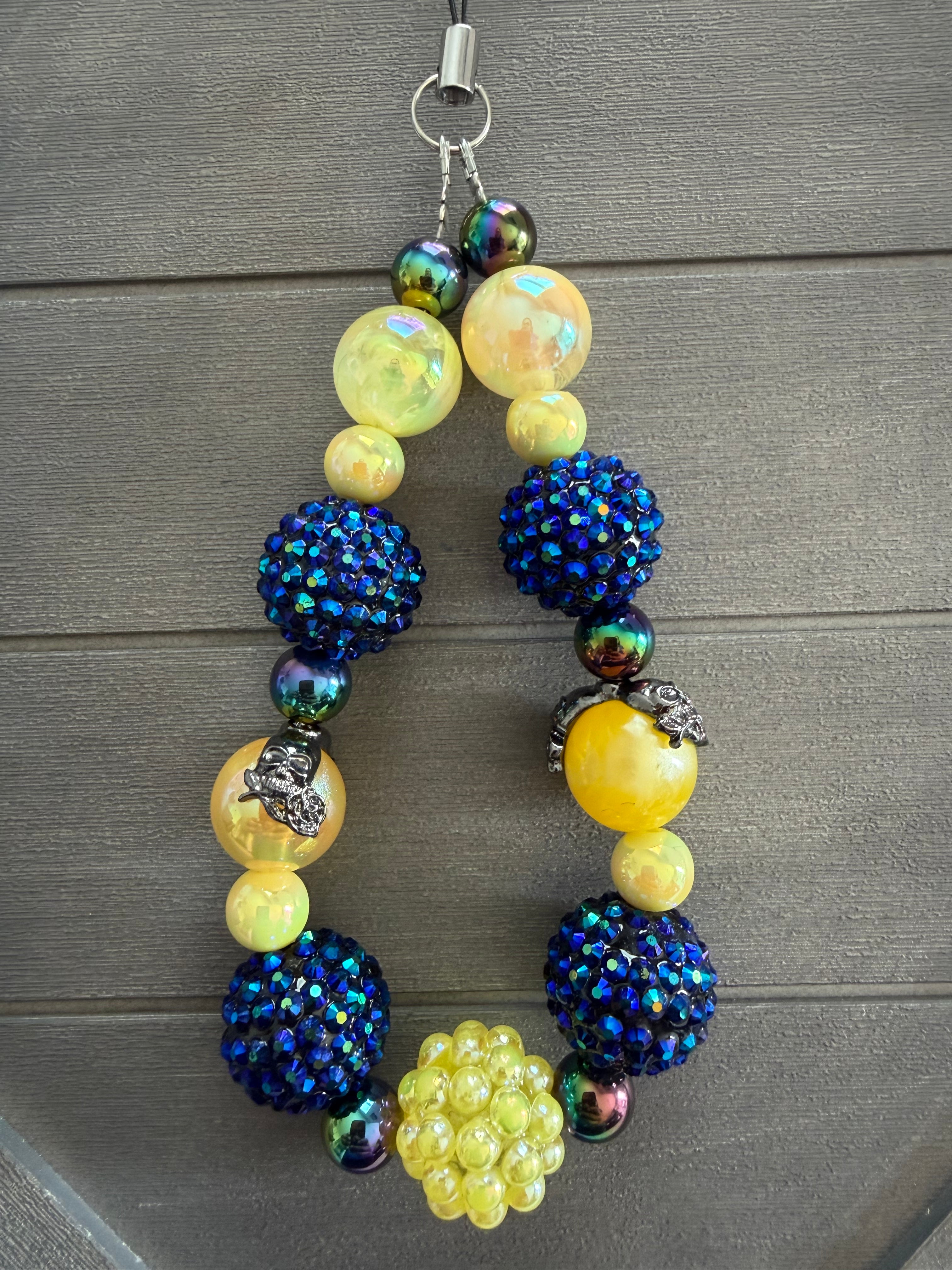 A beaded wristlet with a silver lobster clasp, featuring large iridescent yellow beads, deep blue rhinestone-covered pom-poms, and smaller iridescent beads, displayed on a dark gray wood background.