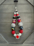 A beaded phone strap or charm with red, black, silver, and white beads, featuring a red rhinestone-encrusted sphere and a central red gemstone in a silver frame, hanging against a dark gray wood background.