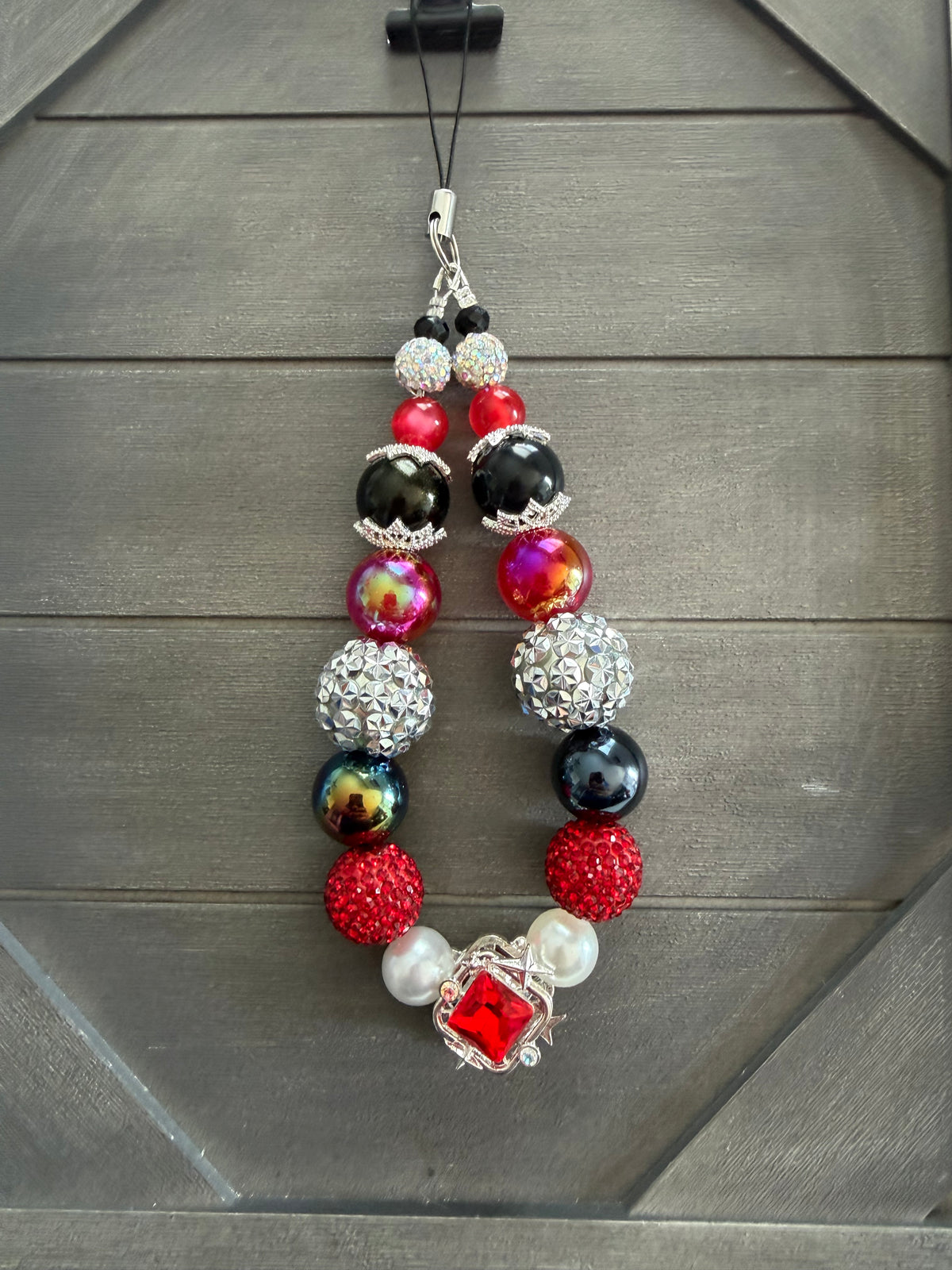 A beaded phone strap or charm with red, black, silver, and white beads, featuring a red rhinestone-encrusted sphere and a central red gemstone in a silver frame, hanging against a dark gray wood background.