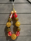 A beaded charm with alternating red translucent beads, gold glittery round beads, and metallic gold pearl-like beads, suspended against a dark gray wood plank background.