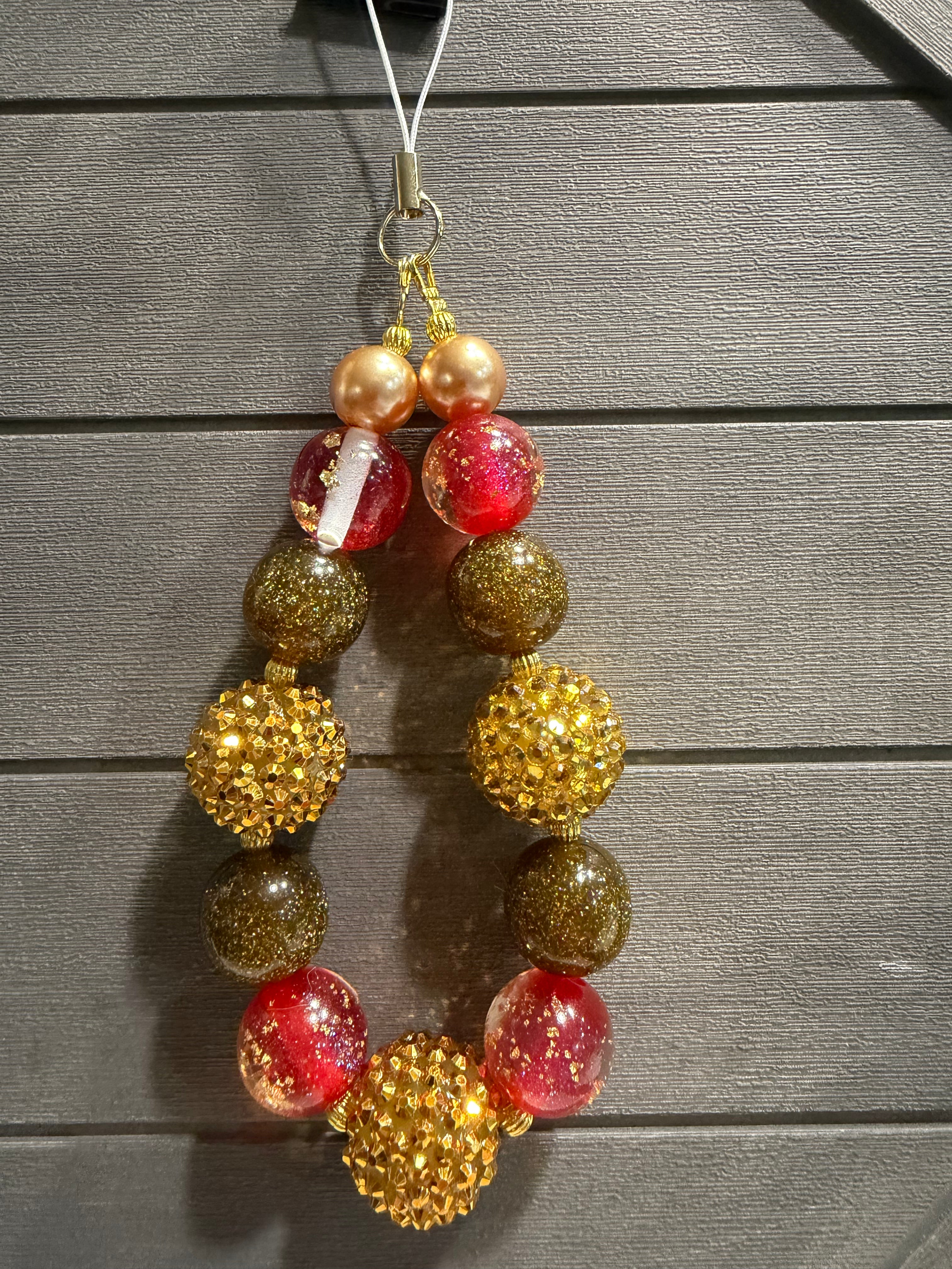 A beaded charm with alternating red translucent beads, gold glittery round beads, and metallic gold pearl-like beads, suspended against a dark gray wood plank background.