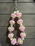 A handmade beaded charm featuring textured white beads with pink hearts and lime green accents, interspersed with translucent purple beads, hanging against a dark gray wooden plank background.