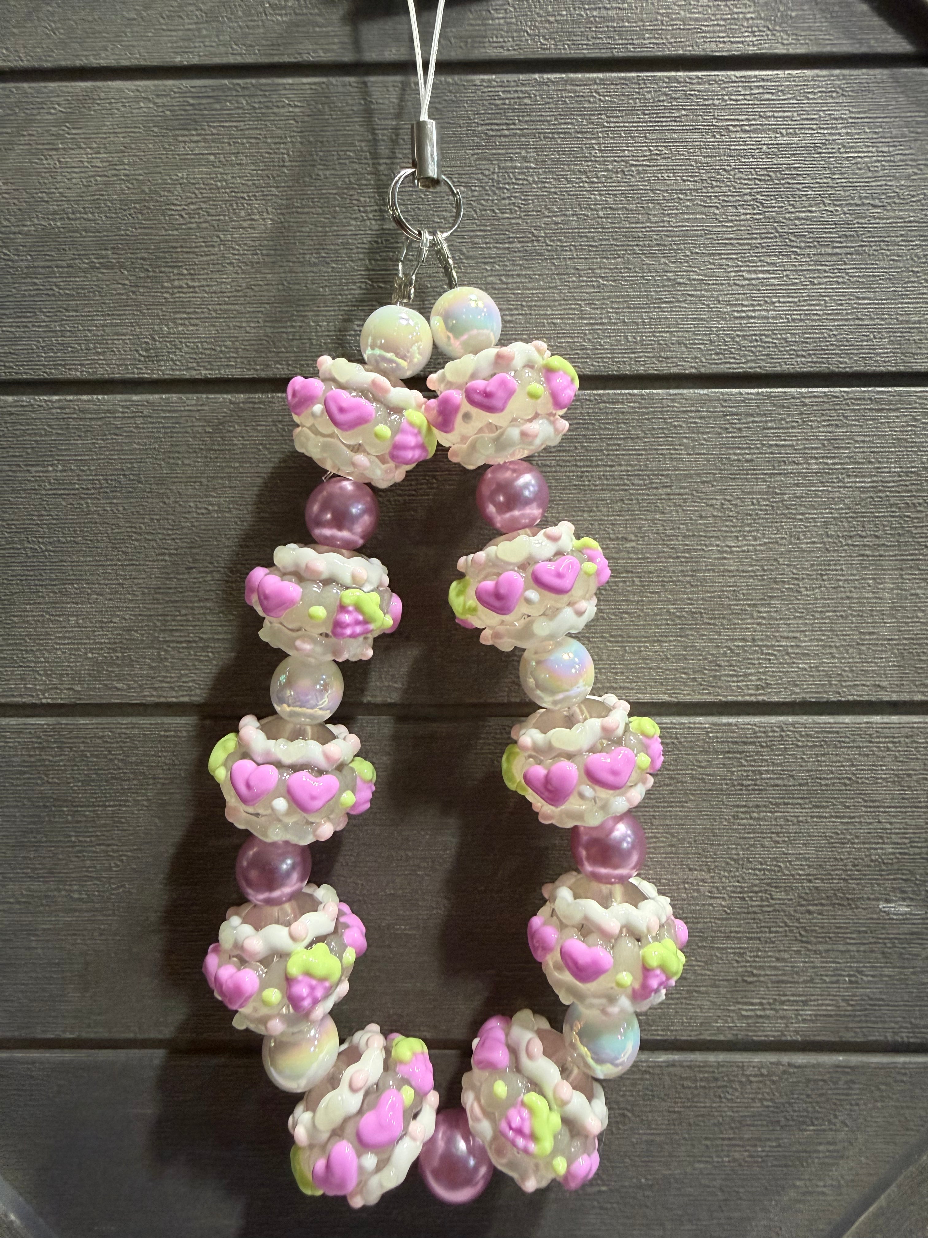 A handmade beaded charm featuring textured white beads with pink hearts and lime green accents, interspersed with translucent purple beads, hanging against a dark gray wooden plank background.