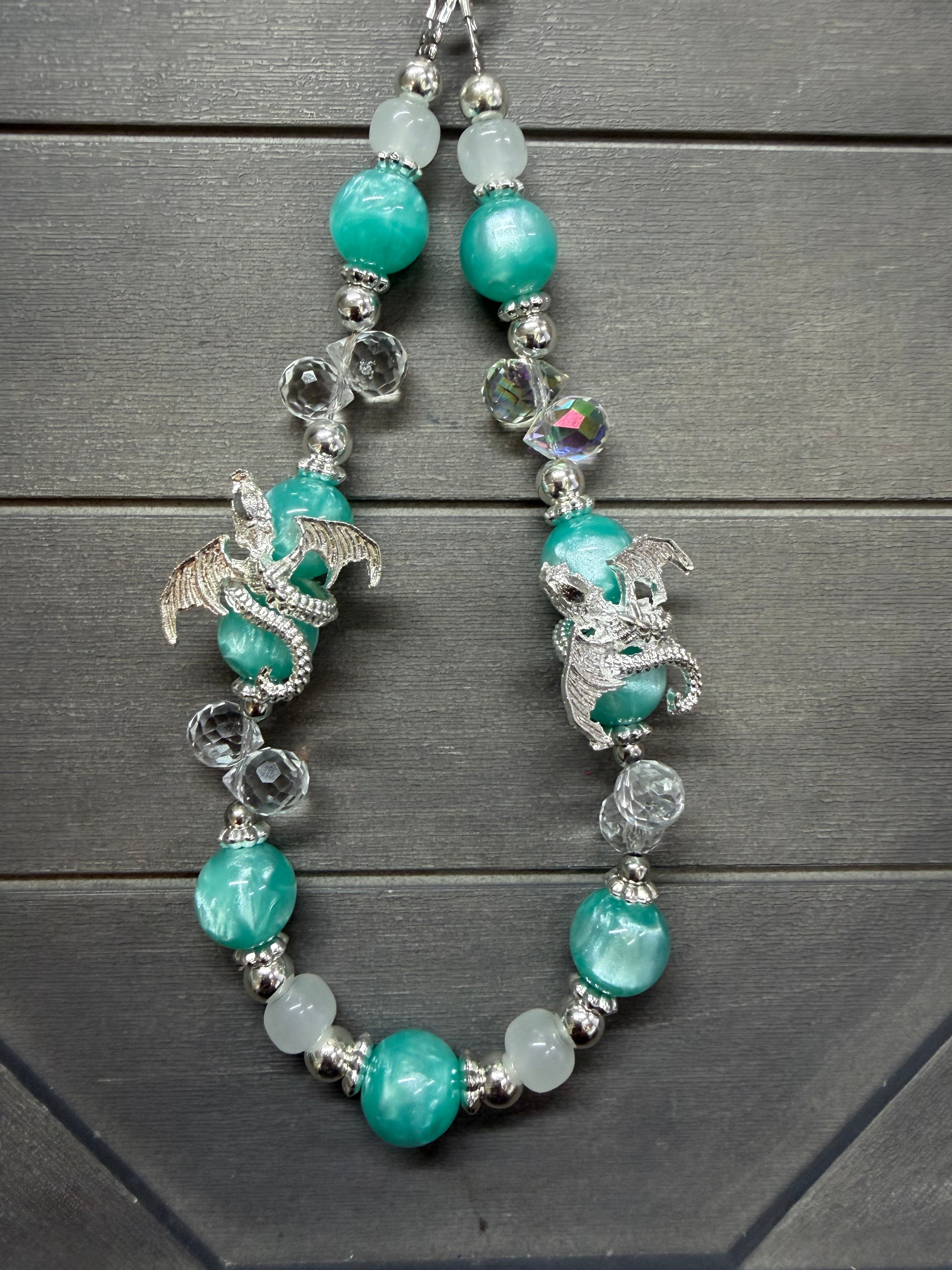  Turquoise and aqua marbled beads, silver dragon charms, clear faceted beads, and iridescent heart beads, displayed on a dark gray wood background.