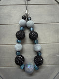 A beaded phone charm with black faceted beads, light gray felt balls, and blue glitter accents, laid flat on a gray wood background.