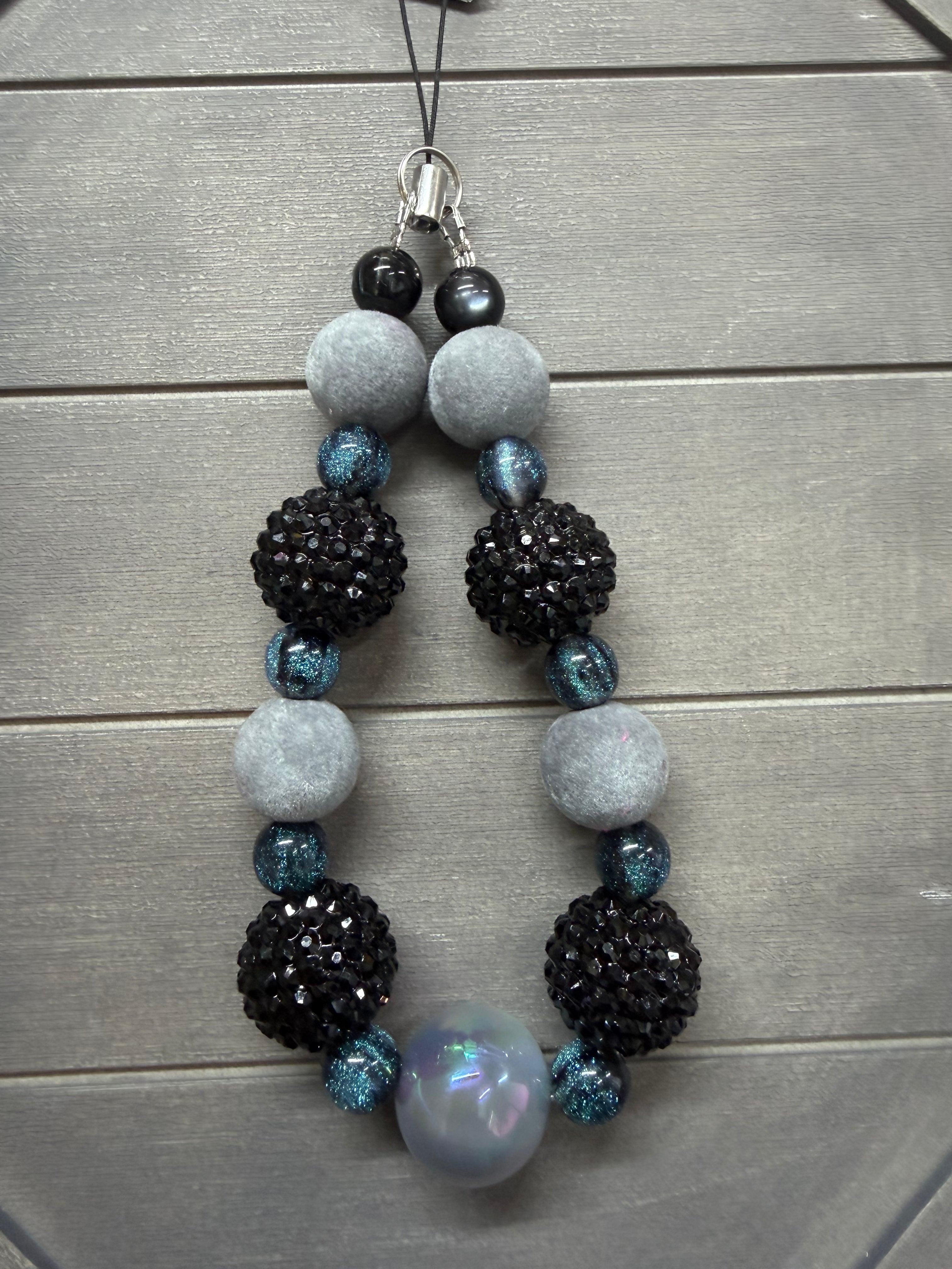 A beaded phone charm with black faceted beads, light gray felt balls, and blue glitter accents, laid flat on a gray wood background.