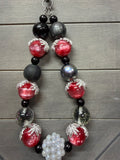 A beaded wristlet featuring large red glass beads, matte black beads, iridescent gray beads, clear beads with glitter, and a cluster of white pearl-like beads, displayed on a gray wood plank background.
