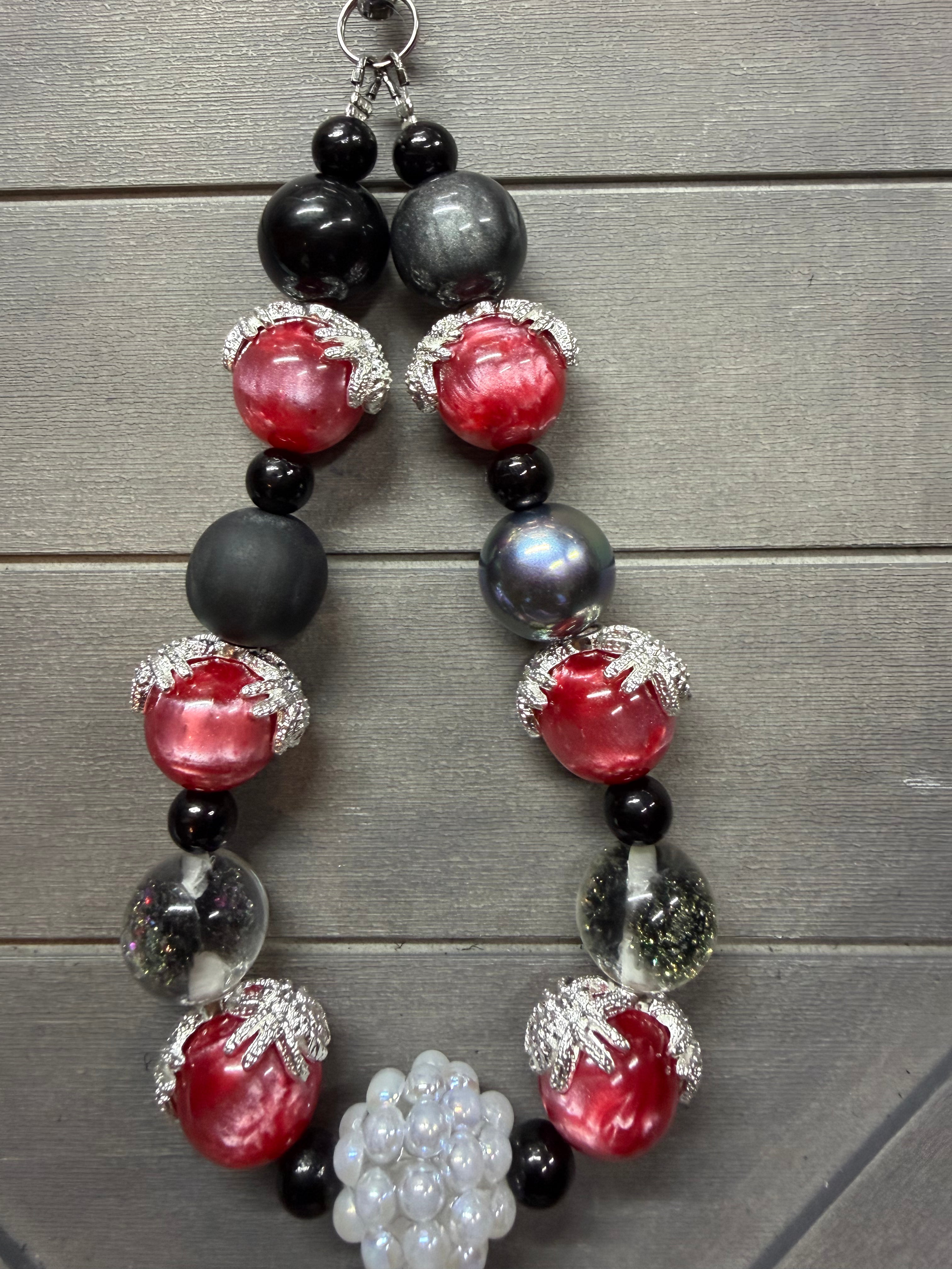 A beaded wristlet featuring large red glass beads, matte black beads, iridescent gray beads, clear beads with glitter, and a cluster of white pearl-like beads, displayed on a gray wood plank background.