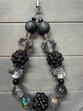 A beaded wristlet featuring a mix of matte black, glossy black, smoky gray, iridescent silver, and holographic beads, displayed on a gray wood plank background.