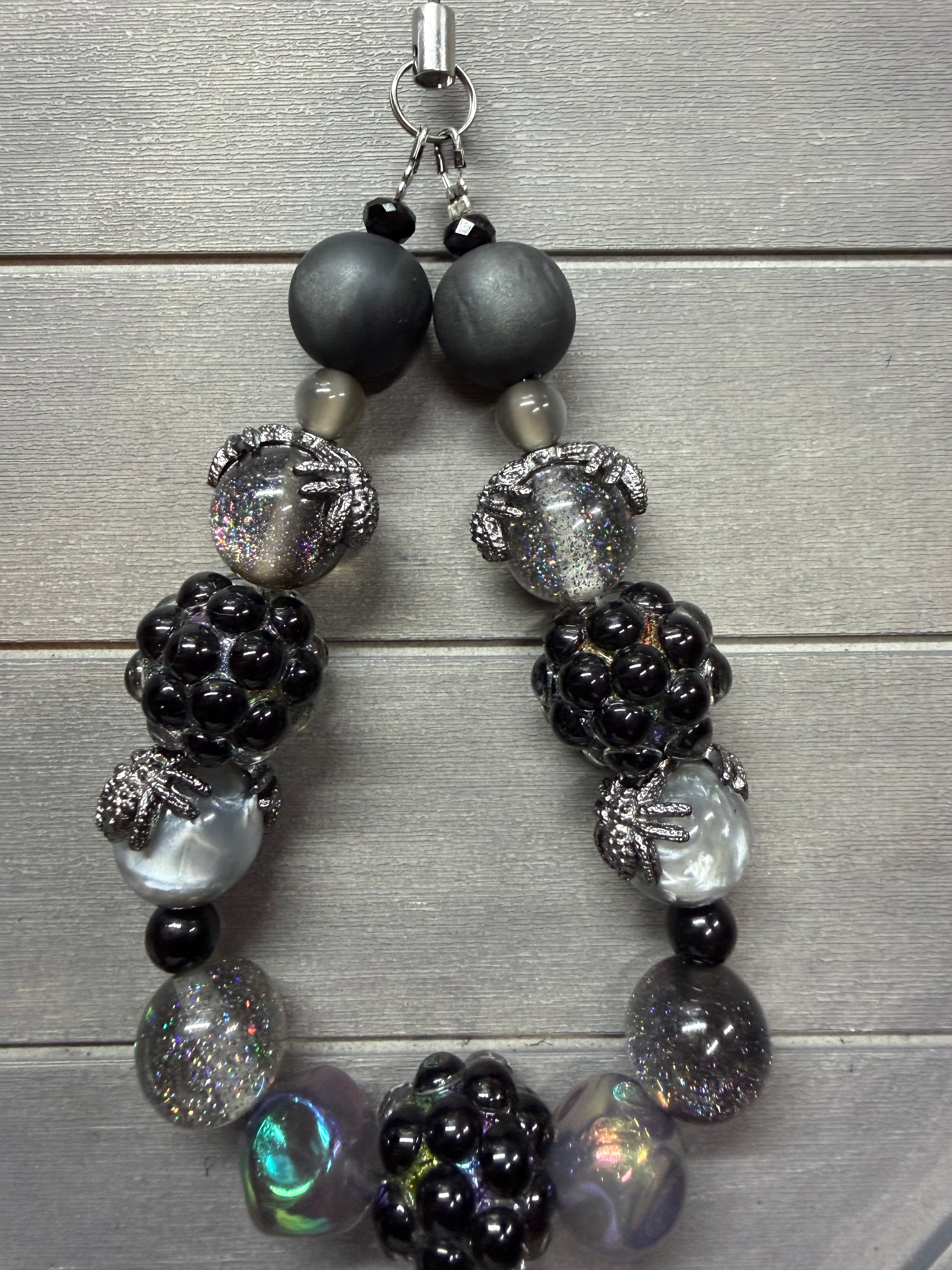A beaded wristlet featuring a mix of matte black, glossy black, smoky gray, iridescent silver, and holographic beads, displayed on a gray wood plank background.