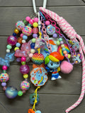 A colorful beaded bag charm with multiple pastel and bright beads, chains, a pink braided strap, a small Rainbow Dash figurine, and cartoon charms, displayed on a dark wood background.