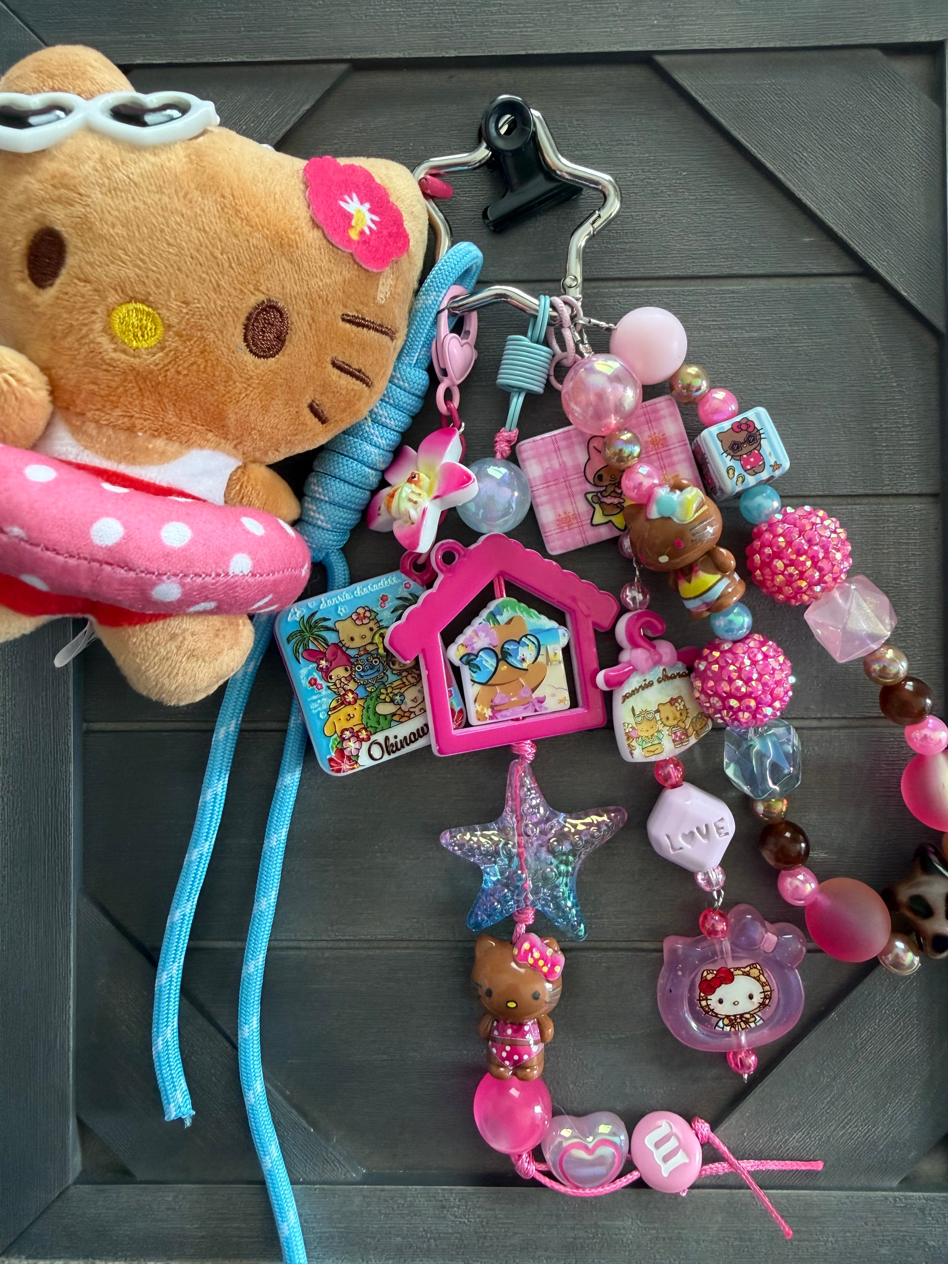 A Hello Kitty themed keychain bundle featuring a plush toy, a pink polka-dot lifebuoy, and multiple colorful charms, beads, and accessories attached to a black binder clip, displayed on a dark gray wooden surface.