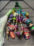 A colorful keychain bundle featuring multiple anime and manga charms, beaded strands, a small character figure, and a green carabiner, displayed on a dark gray wood background.