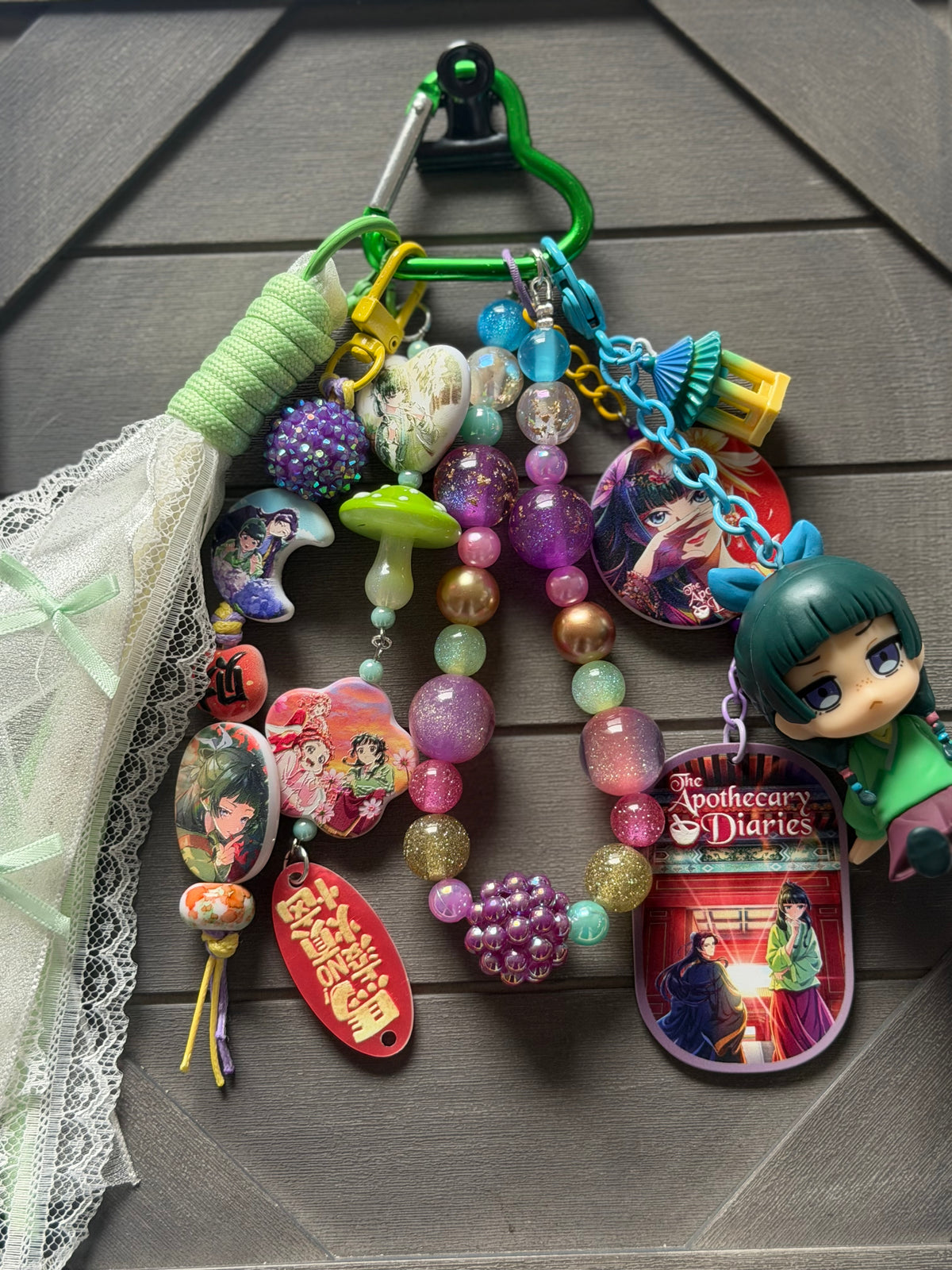 A colorful keychain bundle featuring multiple anime and manga charms, beaded strands, a small character figure, and a green carabiner, displayed on a dark gray wood background.