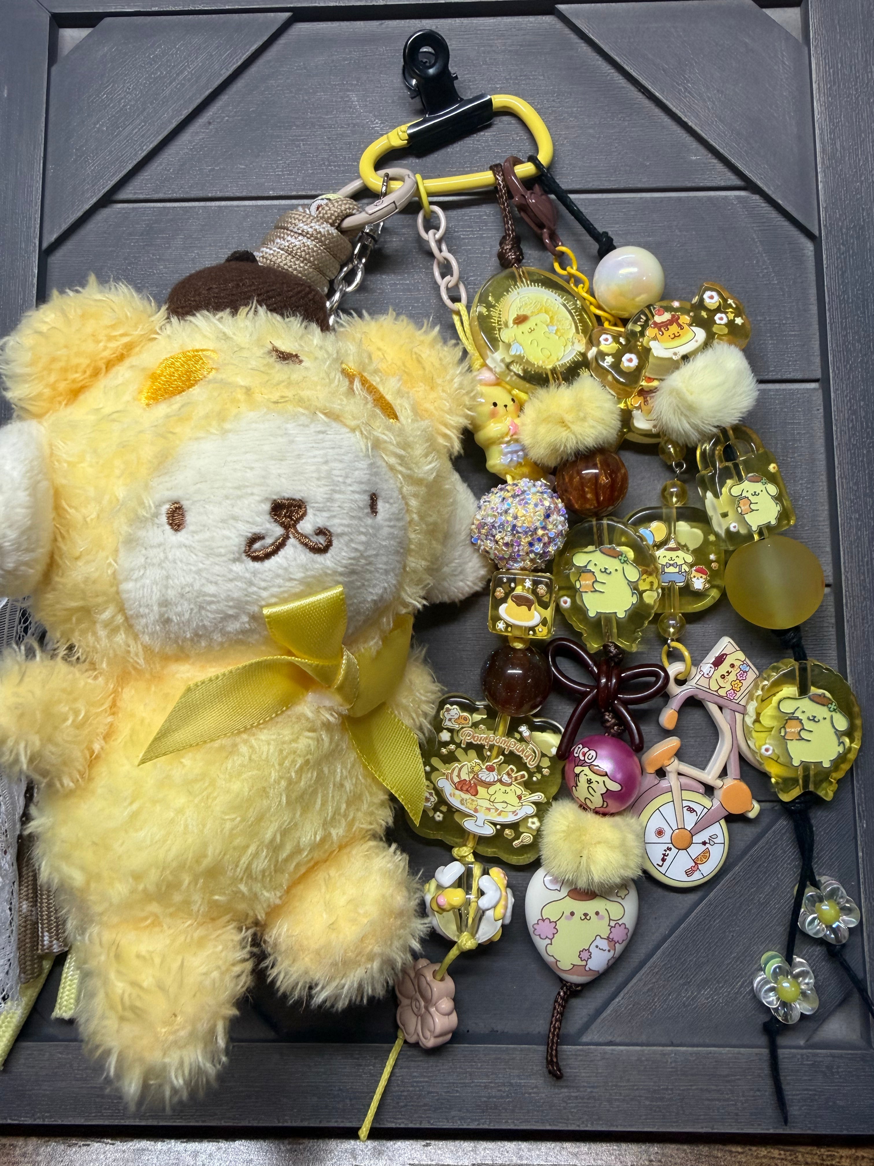 A Purin charm with a matching yellow ribbon, attached to a black carabiner and surrounded by multiple small decorative charms, beads, and acrylic pieces on a dark gray surface.