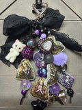 A gothic-themed keychain charm featuring a black lace bow, a small white plush cat, purple and lavender pom-poms, iridescent beads, skull and heart charms, and printed character stickers, displayed on a gray wood background.