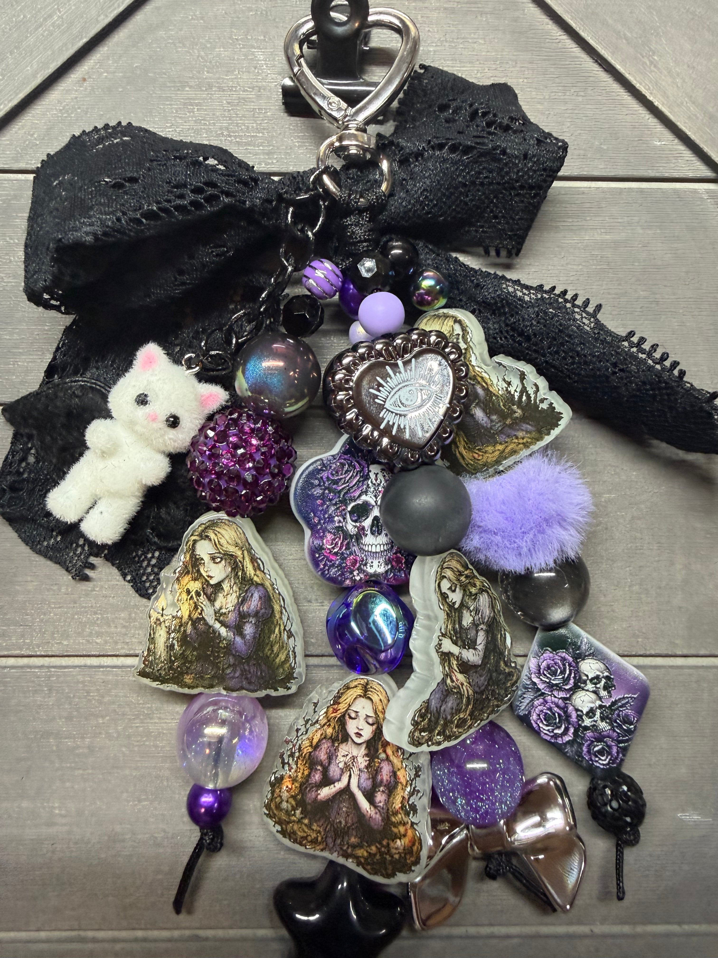 A gothic-themed keychain charm featuring a black lace bow, a small white plush cat, purple and lavender pom-poms, iridescent beads, skull and heart charms, and printed character stickers, displayed on a gray wood background.