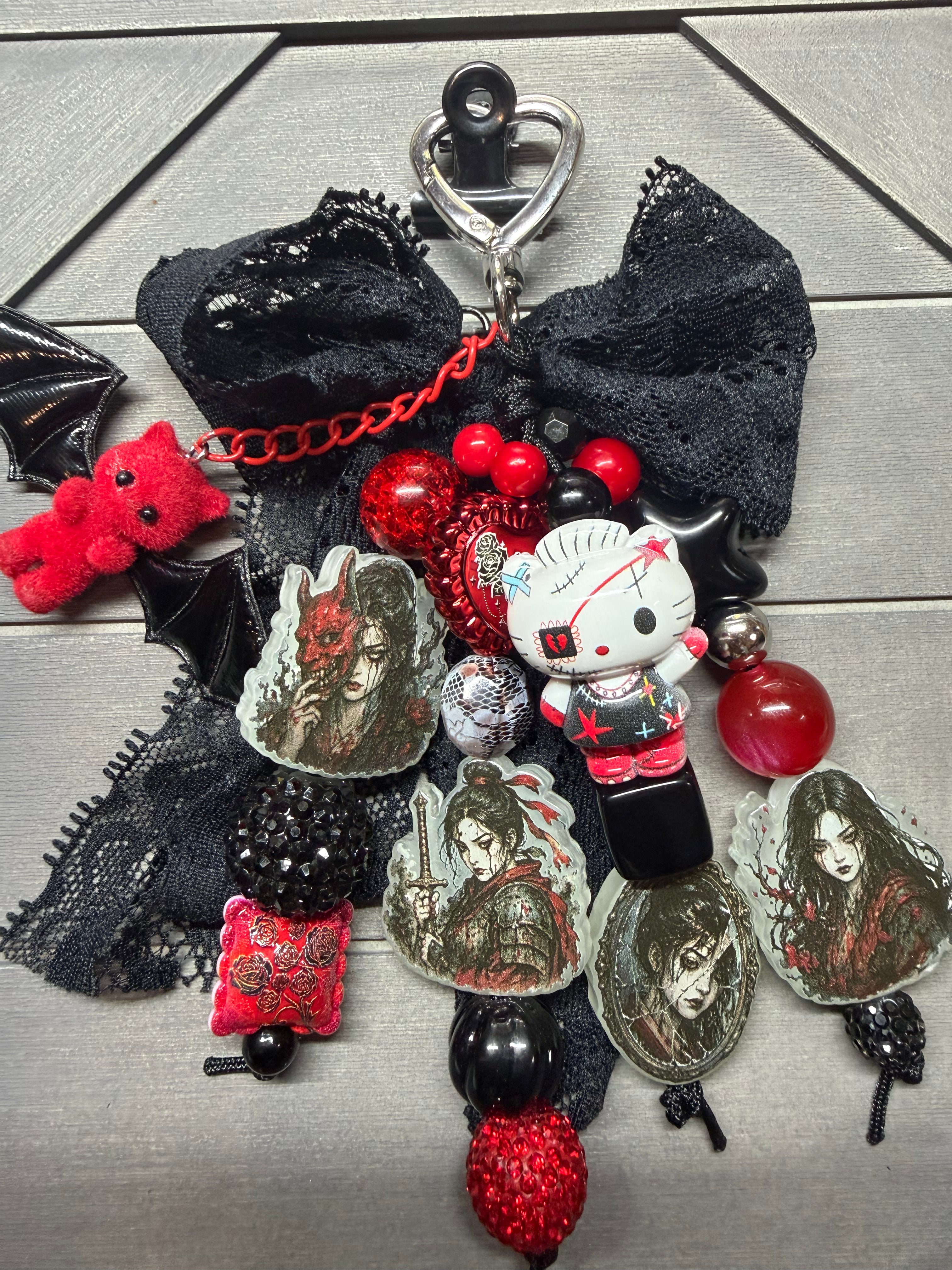 A gothic-themed bag charm featuring a large black lace bow, red beads, a small Hello Kitty figure, bat wings, and multiple character portrait charms, displayed on a gray wood background.