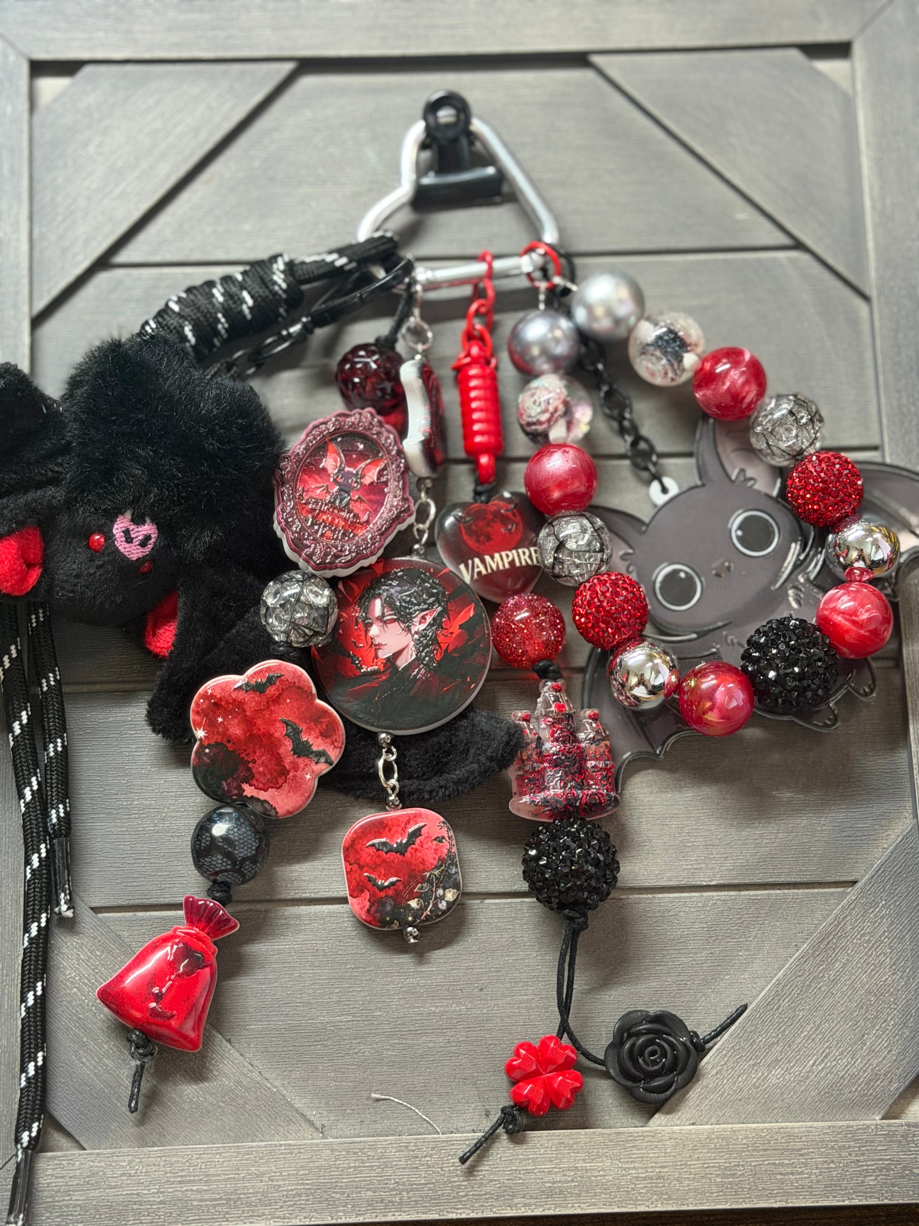 A gothic-themed  bag charm set featuring a black plush bat, a gray bat figure, red and black beads, bat-shaped charms, a small red bell, and a black rose, displayed on a gray wood-grain surface.