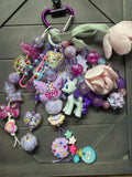 A colorful My Little Pony themed keychain bag charm set featuring a purple carabiner, a mint green ribbon, a pink plush flower, a purple rose, a pony figure, and various pastel charms on a dark gray wood background.