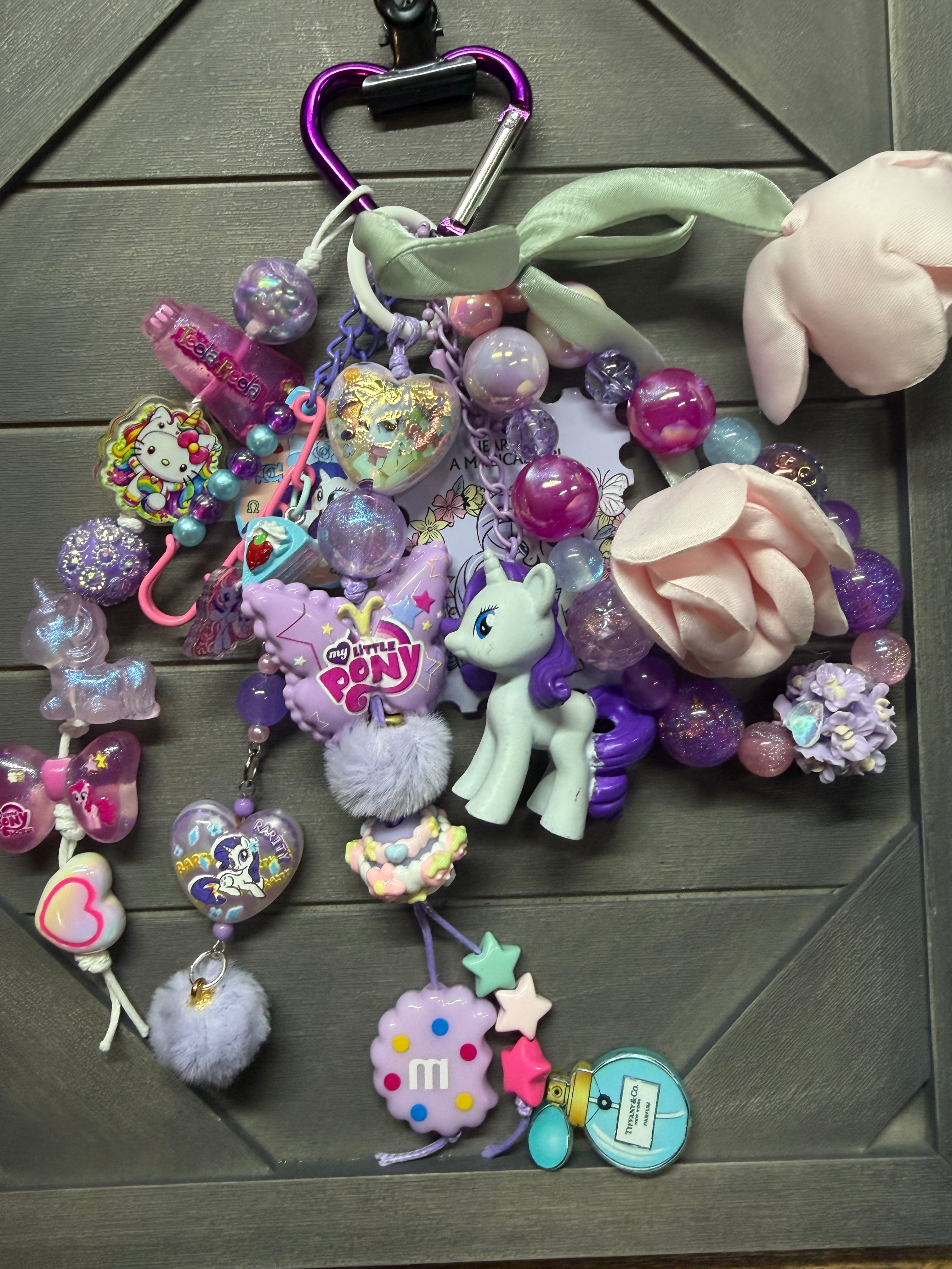 A colorful My Little Pony themed keychain bag charm set featuring a purple carabiner, a mint green ribbon, a pink plush flower, a purple rose, a pony figure, and various pastel charms on a dark gray wood background.