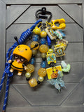 A colorful beaded keychain or bag charm set with a black metal clasp, featuring yellow and blue beads, a small bee character figure, a blue rope, a butterfly charm, and multiple decorative tags, displayed on a dark gray paneled surface.