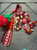 A strawberry-themed beaded bag charm set displayed on a dark gray wooden surface. The set includes a large red and green crocheted strawberry, red and pink translucent beads, small strawberry-shaped beads, a floral bead, a red-and-white gingham ribbon, and a black plastic carabiner clip.