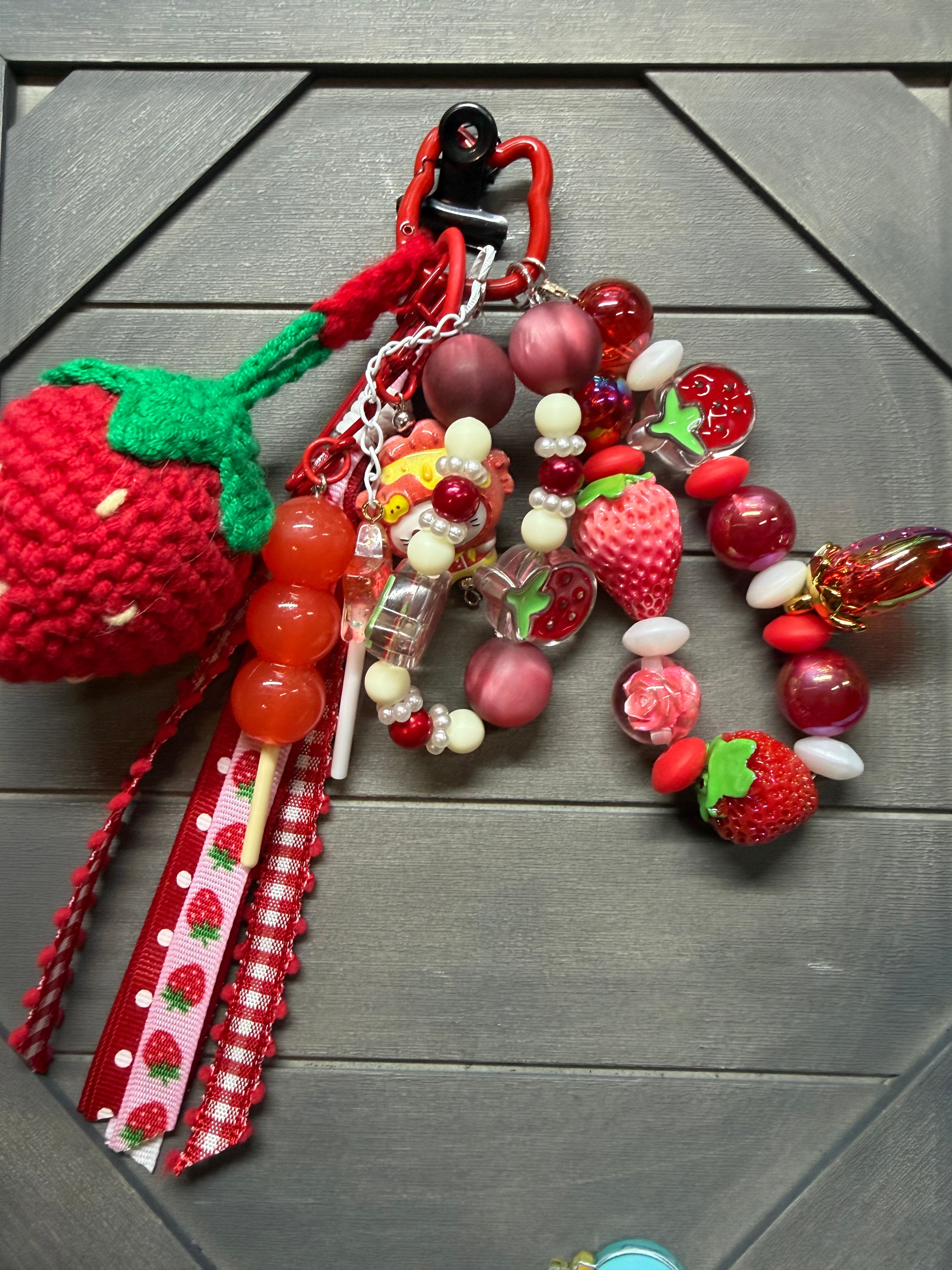 A strawberry-themed beaded bag charm set displayed on a dark gray wooden surface. The set includes a large red and green crocheted strawberry, red and pink translucent beads, small strawberry-shaped beads, a floral bead, a red-and-white gingham ribbon, and a black plastic carabiner clip.