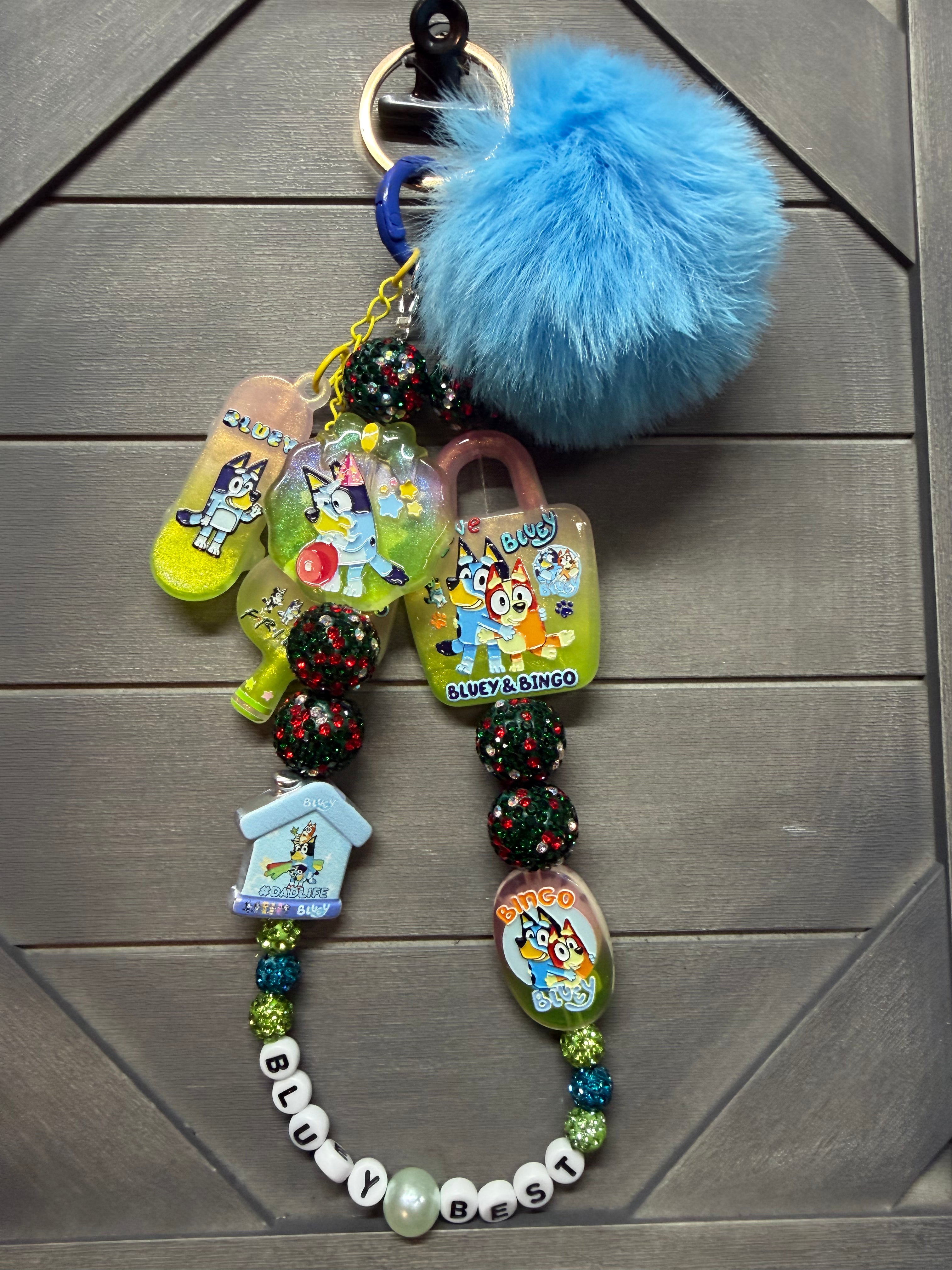 A colorful custom keychain featuring cartoon character charms, a blue pom-pom, and Bluey on a dark gray paneled background.